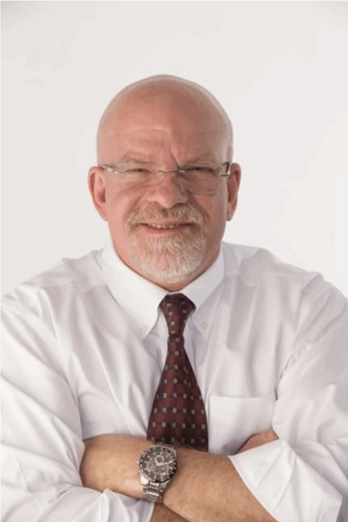 Bald man with a white beard and a white shirt holds arms crossed over his chest and smiles at the camera